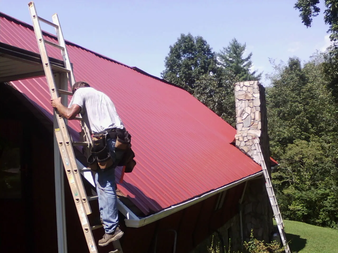 Roofer on a ladder beside a red metal roof and stone chimney — Soffit & Fascia Repair by Roofing Brothers in Hemet